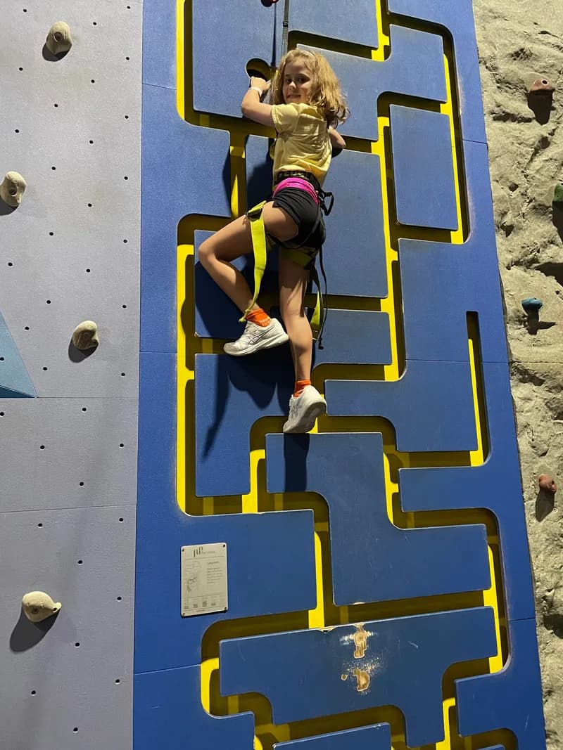 A child, wearing a safety harness, pausing to look at the viewer, while halfway up an indoor rock climbing wall.